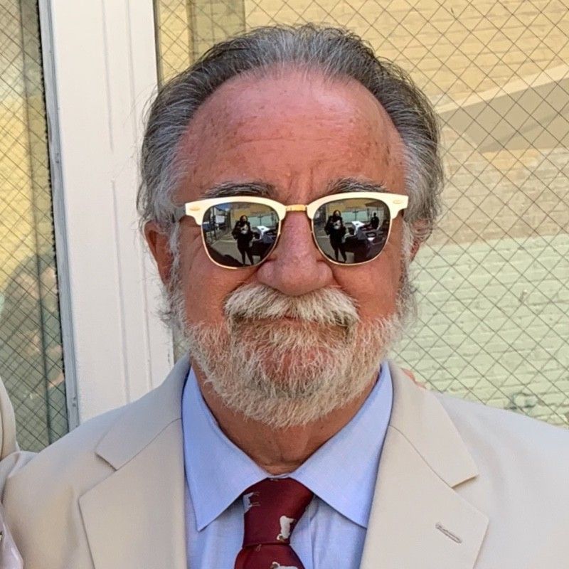 Man in beige suit and red tie wearing mirrored sunglasses, standing outdoors by a wire-mesh door.