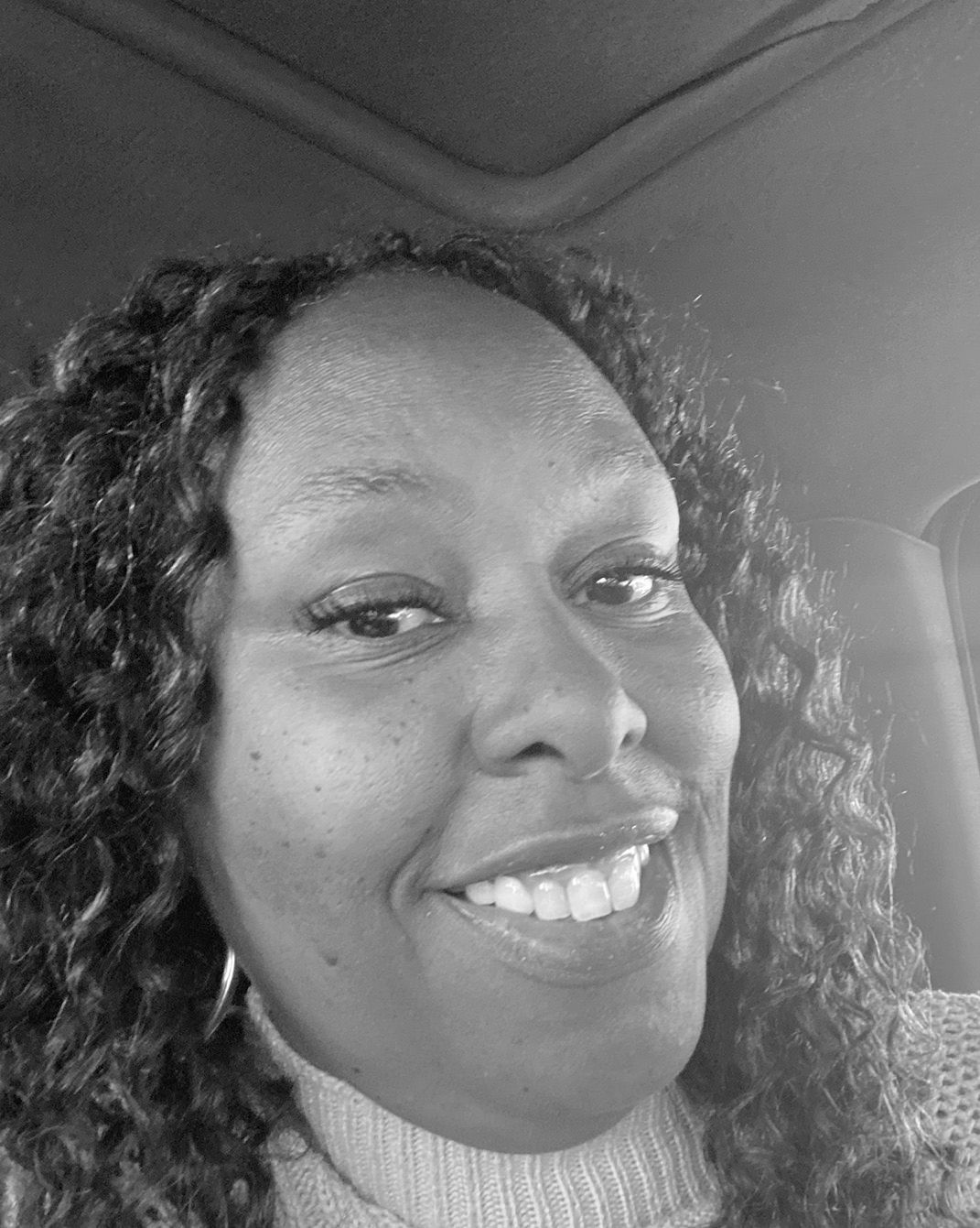 Smiling person in a car, wearing a light turtleneck sweater, in a black-and-white selfie.