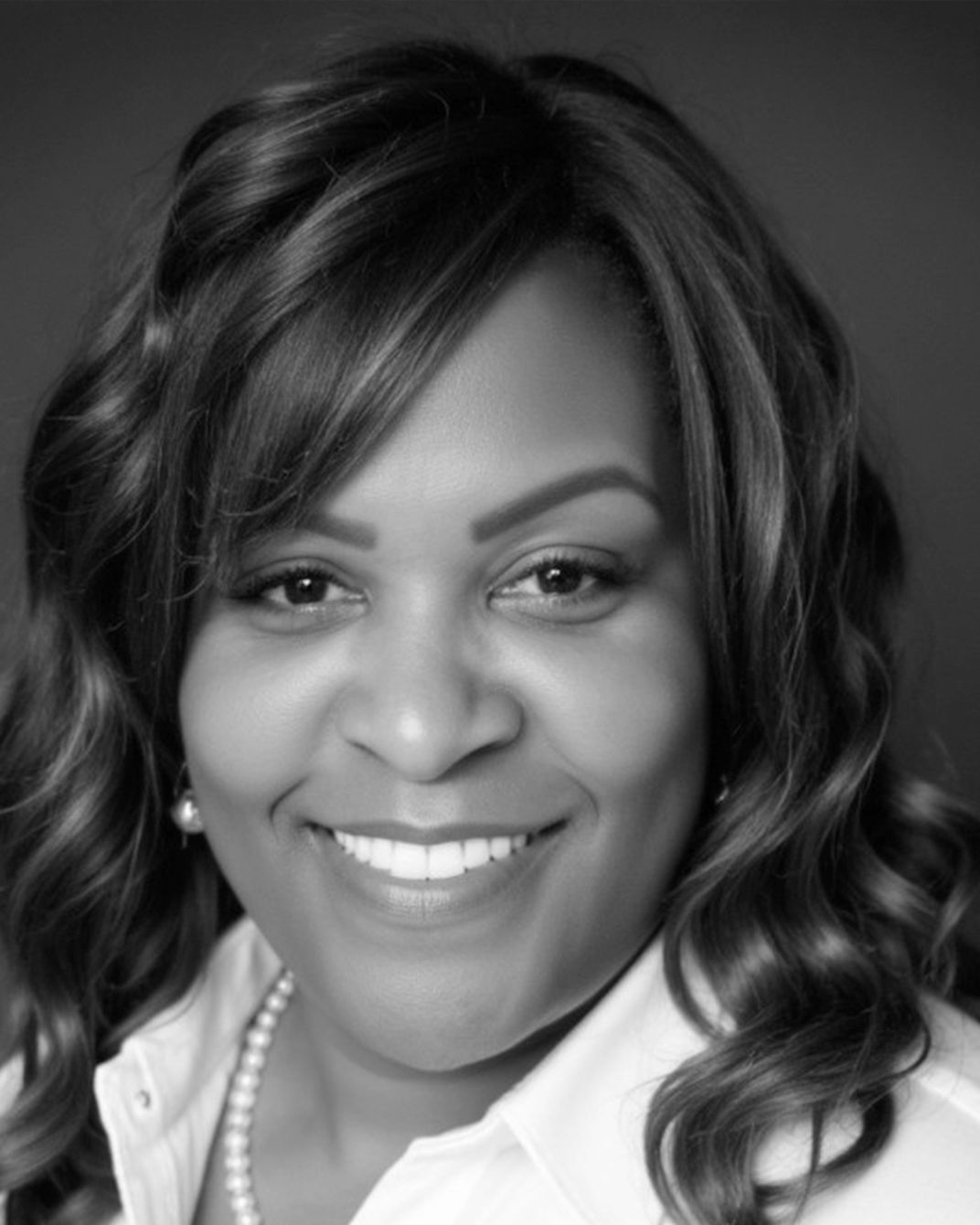 Smiling woman with wavy hair, wearing a white blouse and pearl necklace, against a dark background