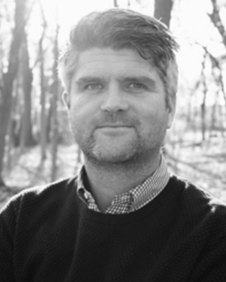 Portrait of a man outdoors in a black sweater, standing before bare trees in soft light