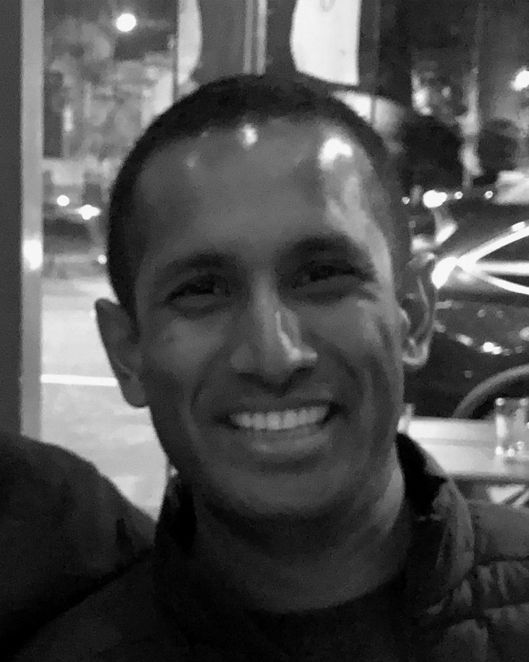 Smiling man in a dark jacket at night, with city lights and parked cars in the background.