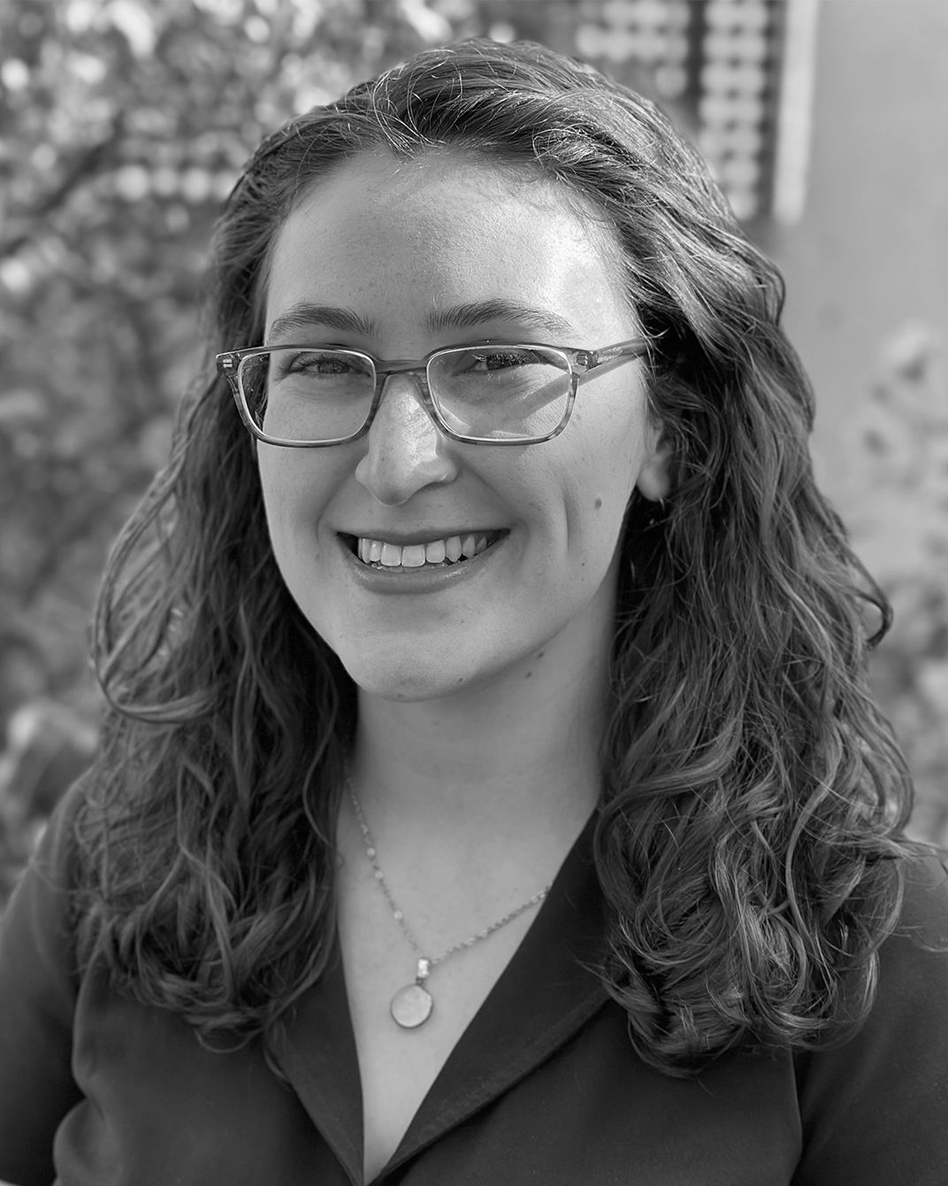 Smiling person with glasses and curly hair, wearing a dark shirt and pendant necklace outdoors.