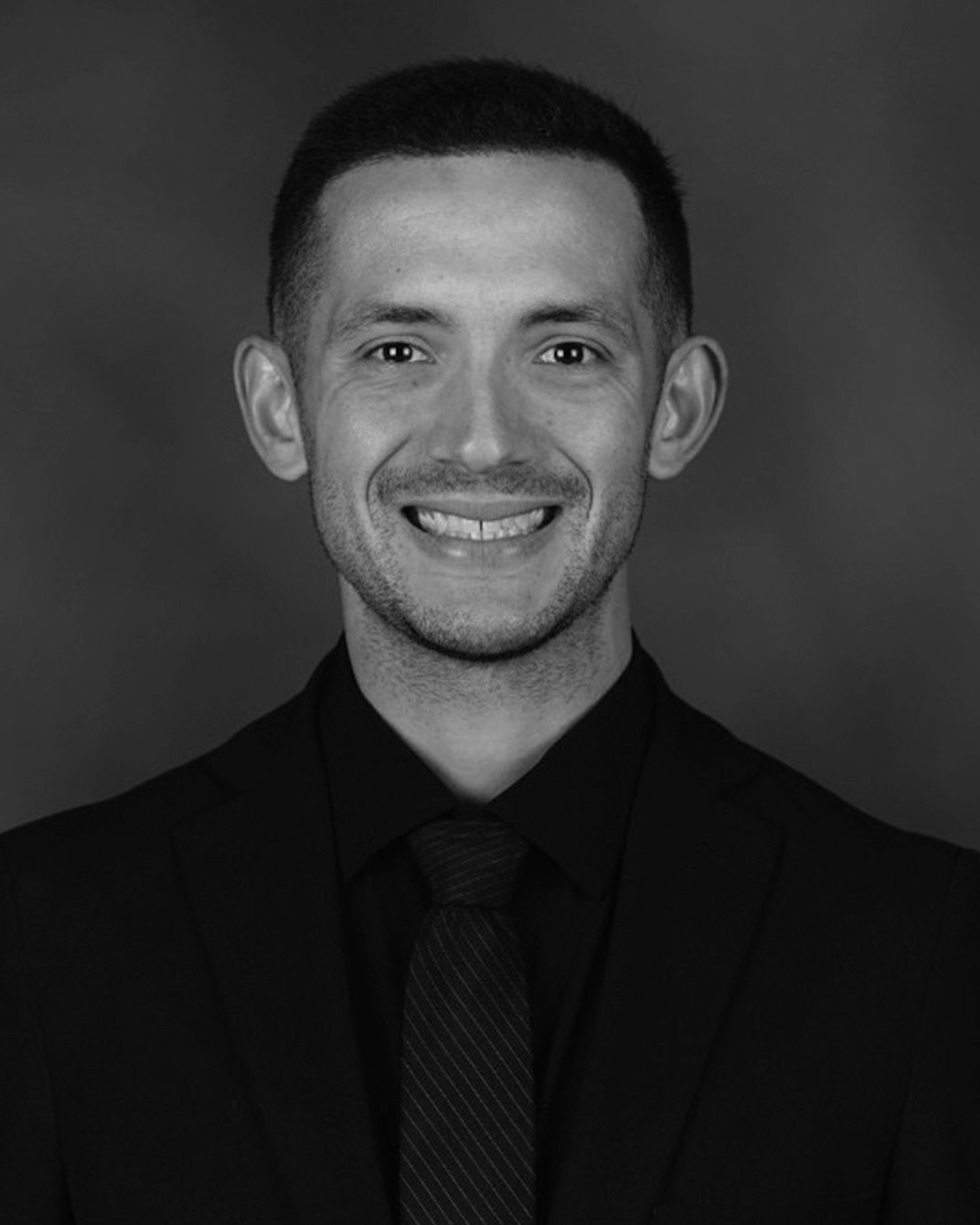 Studio portrait of a smiling man in a dark suit and tie against a plain gray background