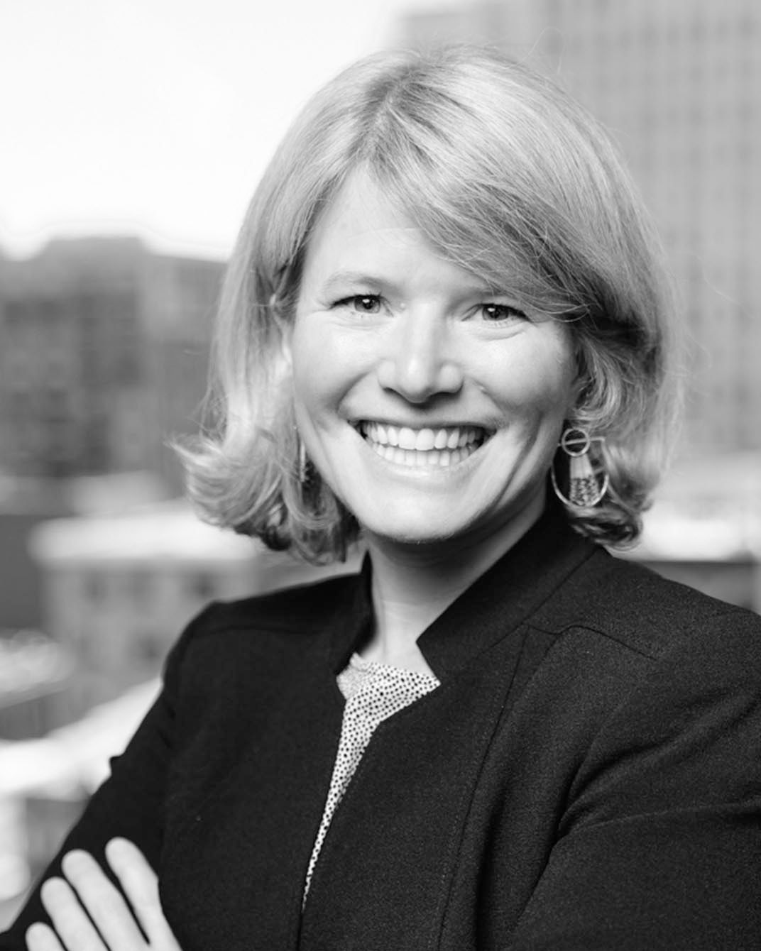 Smiling woman in a dark blazer with arms crossed, standing before a blurred city background