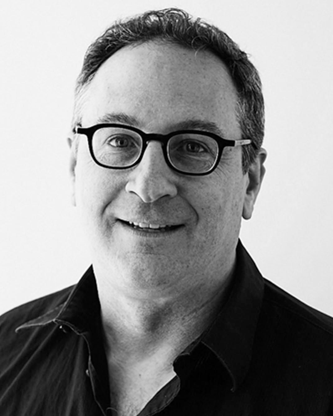 Black-and-white headshot of a smiling man wearing glasses and a dark shirt