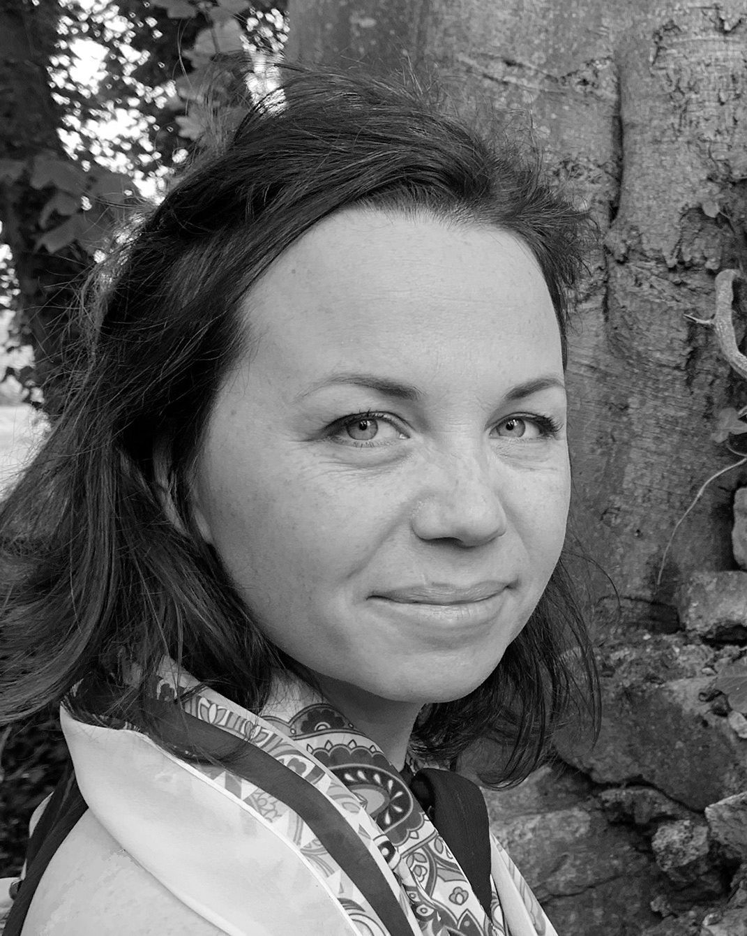 Black-and-white portrait of a woman standing by a tree, looking at the camera with a slight smile.