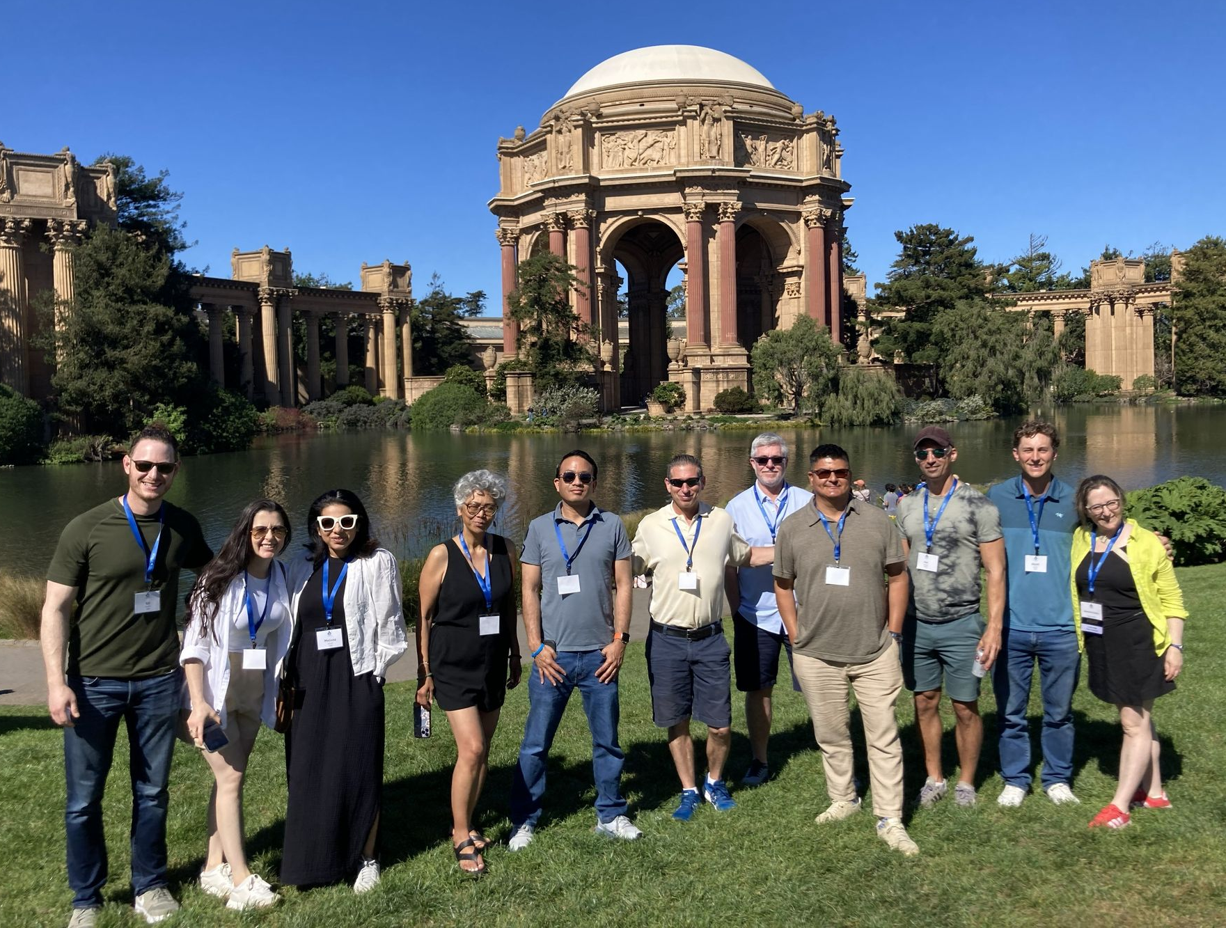 Group of people posing on a grassy lawn in front of a domed classical ruin under a blue sky