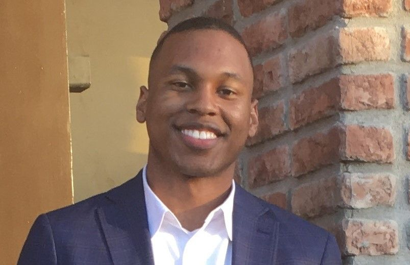 Smiling person in a navy blazer and white shirt standing by a brick wall and doorway