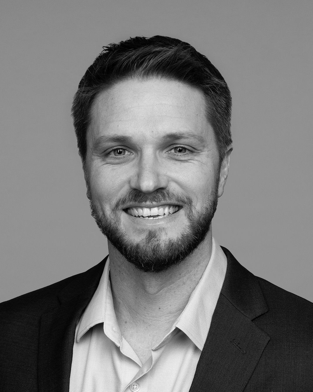 Smiling man in a dark blazer and light shirt against a plain gray background