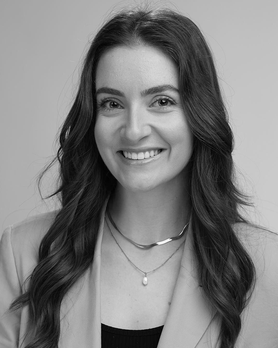 Smiling woman with long dark hair wearing layered necklaces against a light background