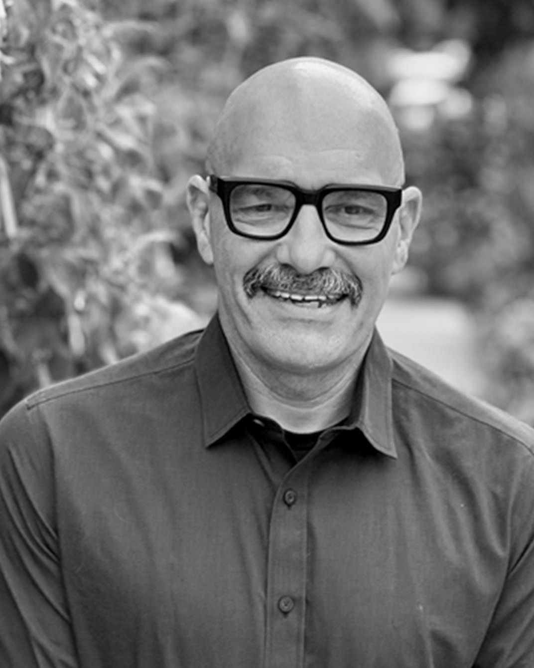 Portrait of a smiling man wearing glasses and a dark shirt, posed outdoors with a blurred background