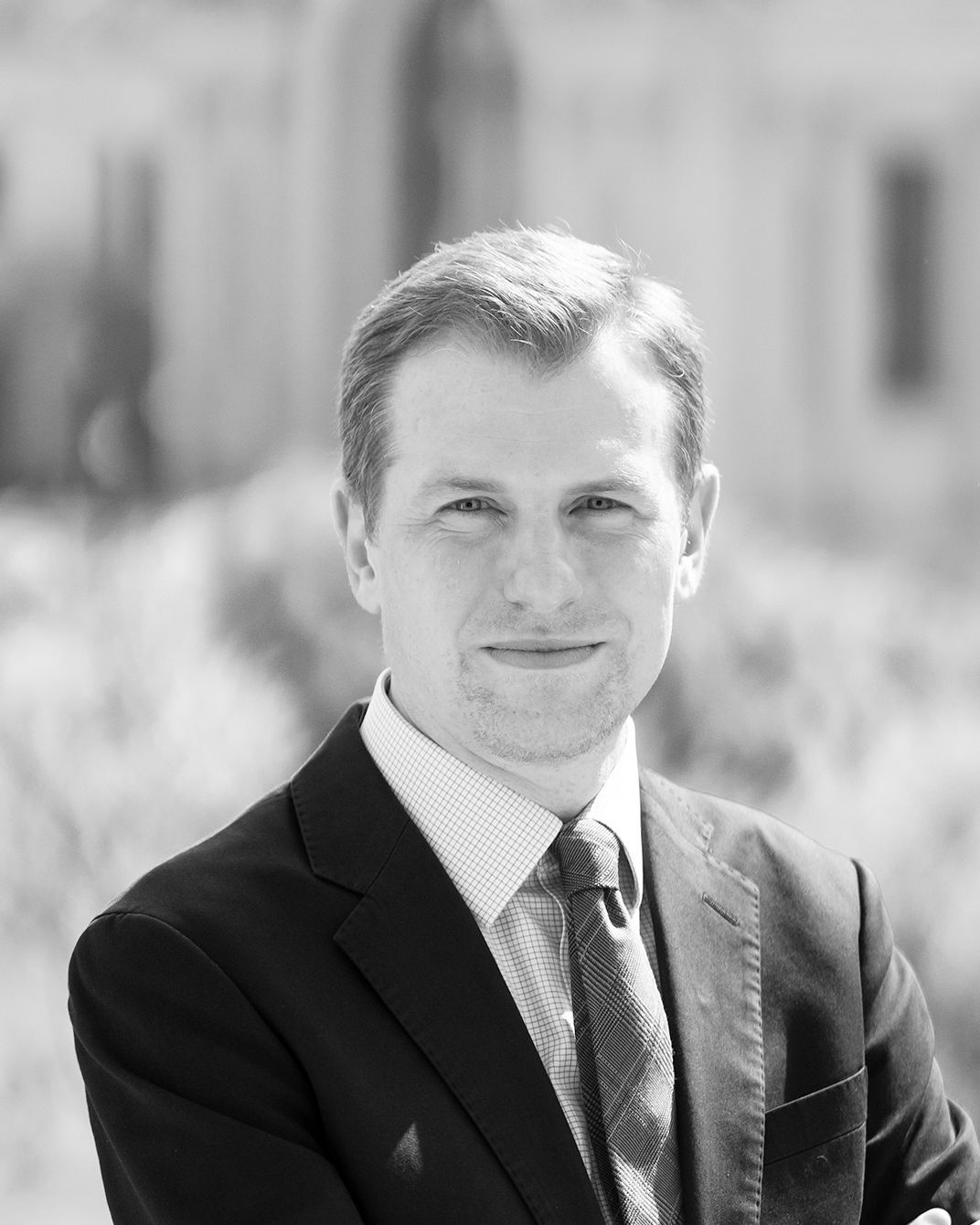 Black-and-white portrait of a man in a suit and tie, standing outdoors with a blurred building background