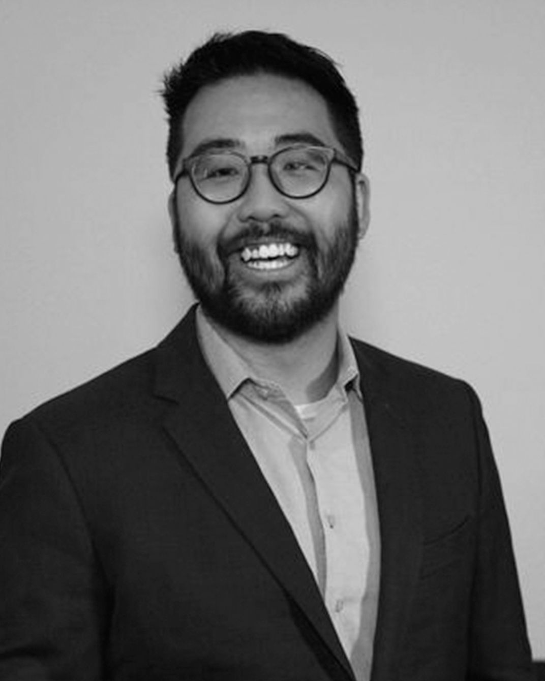 Smiling man in glasses and a dark suit jacket, posing against a plain wall.