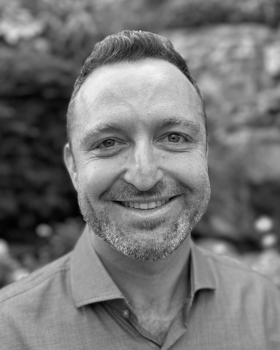 Smiling man in a collared shirt, photographed outdoors in black and white.