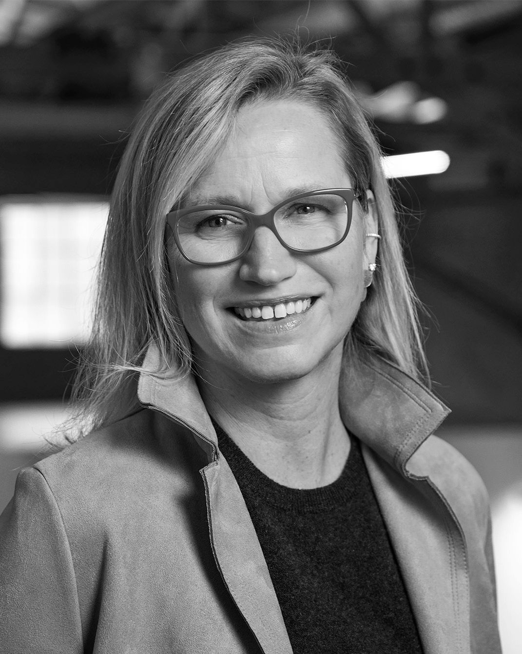 Smiling woman with glasses in a blazer, standing indoors in a black-and-white portrait