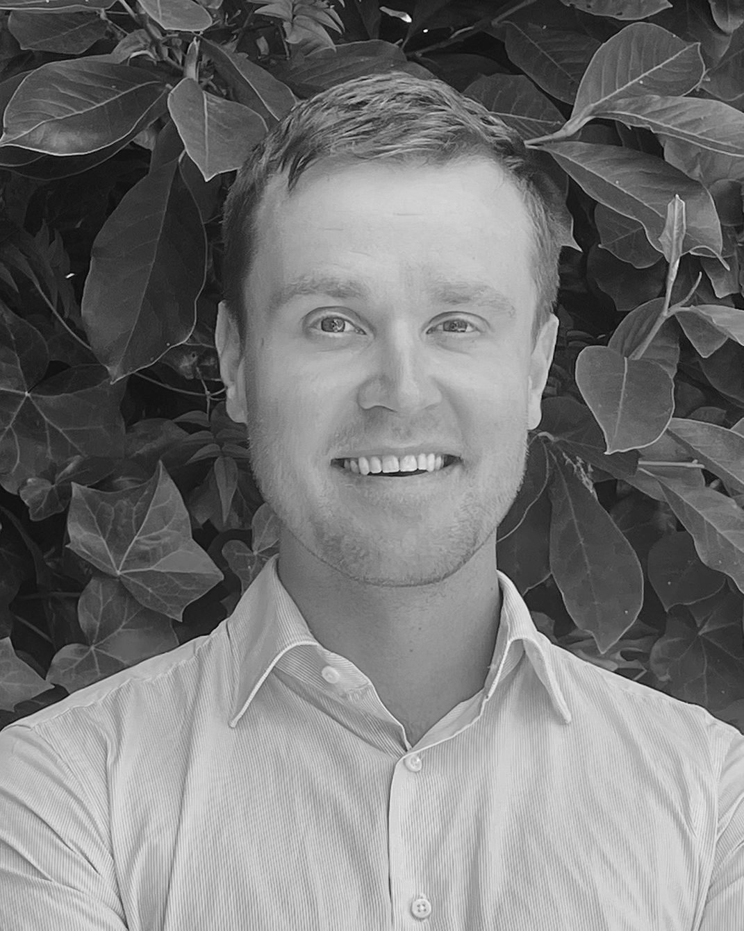 Smiling person in a collared shirt standing in front of leafy foliage.