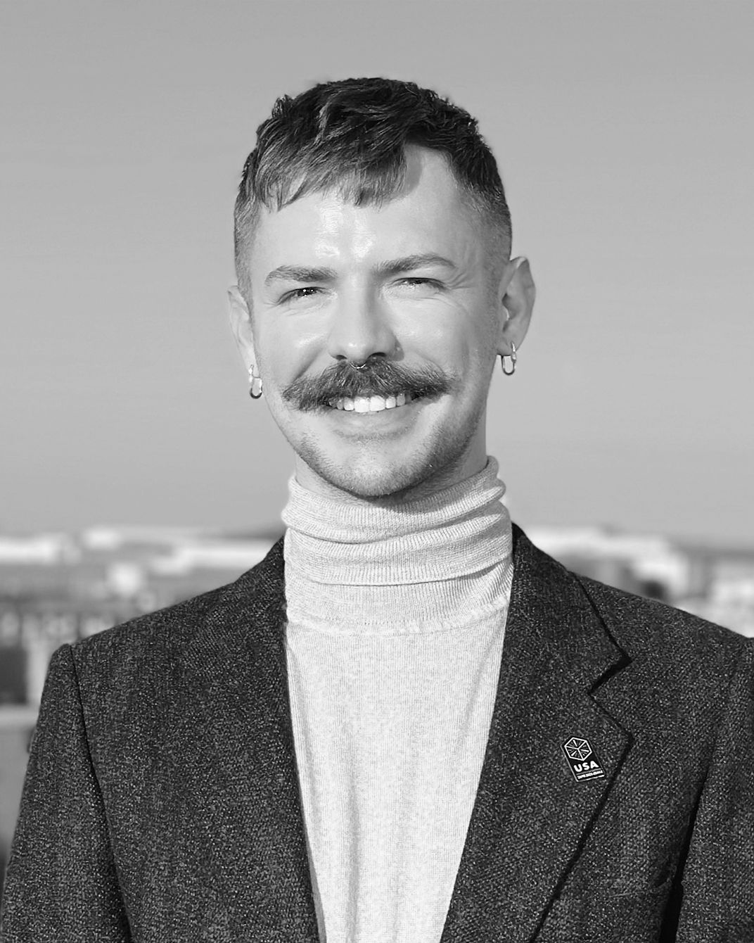Portrait of a smiling person with a mustache, wearing a turtleneck and blazer, against a blurred outdoor background.