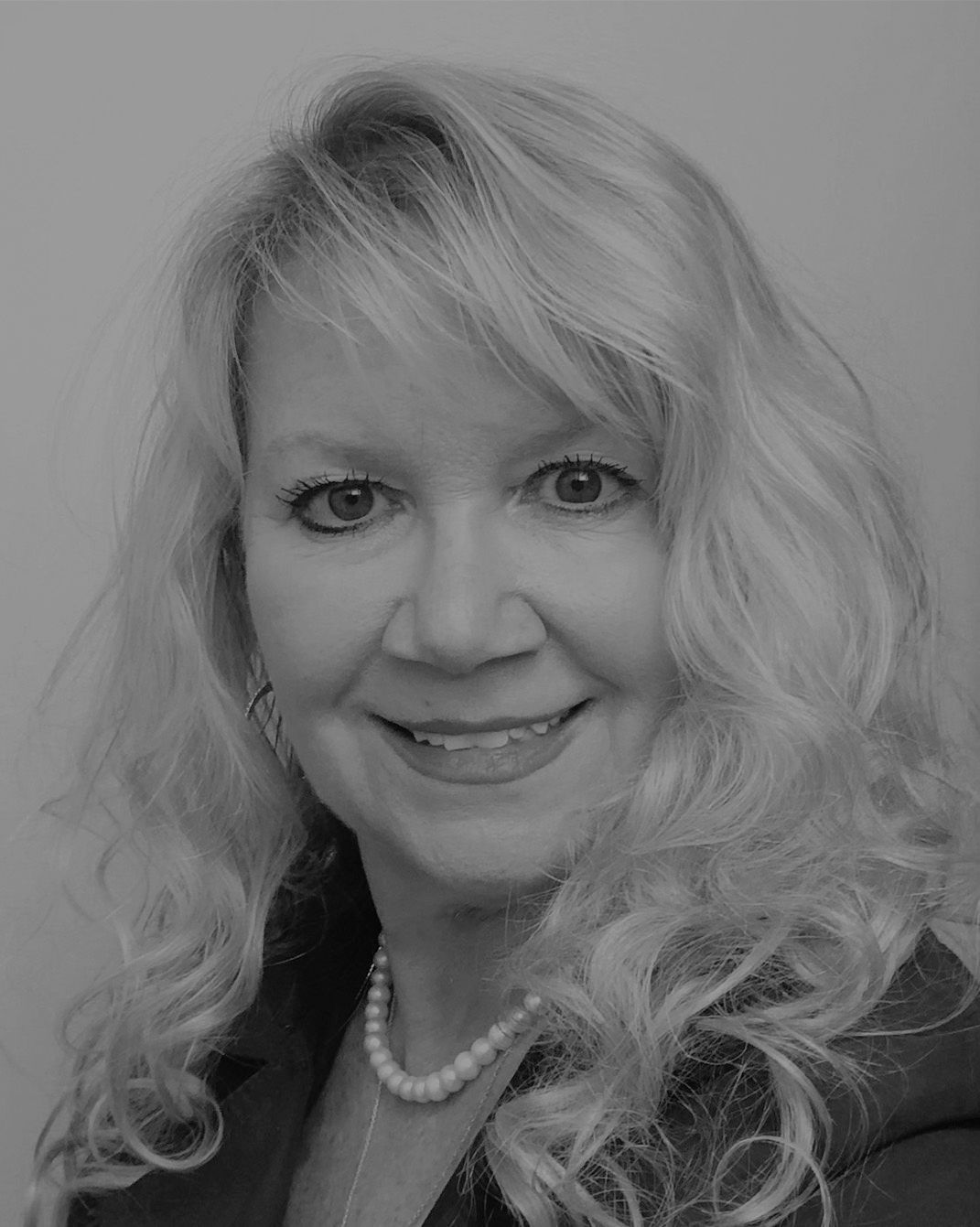 Smiling woman with wavy blonde hair, wearing a pearl necklace and dark jacket.