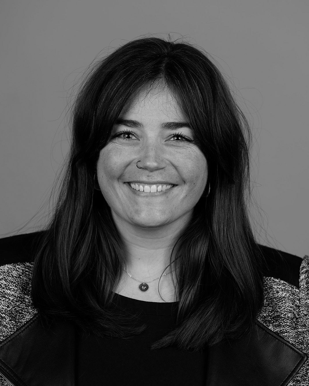 Smiling woman with long dark hair, wearing a patterned jacket, against a plain gray background