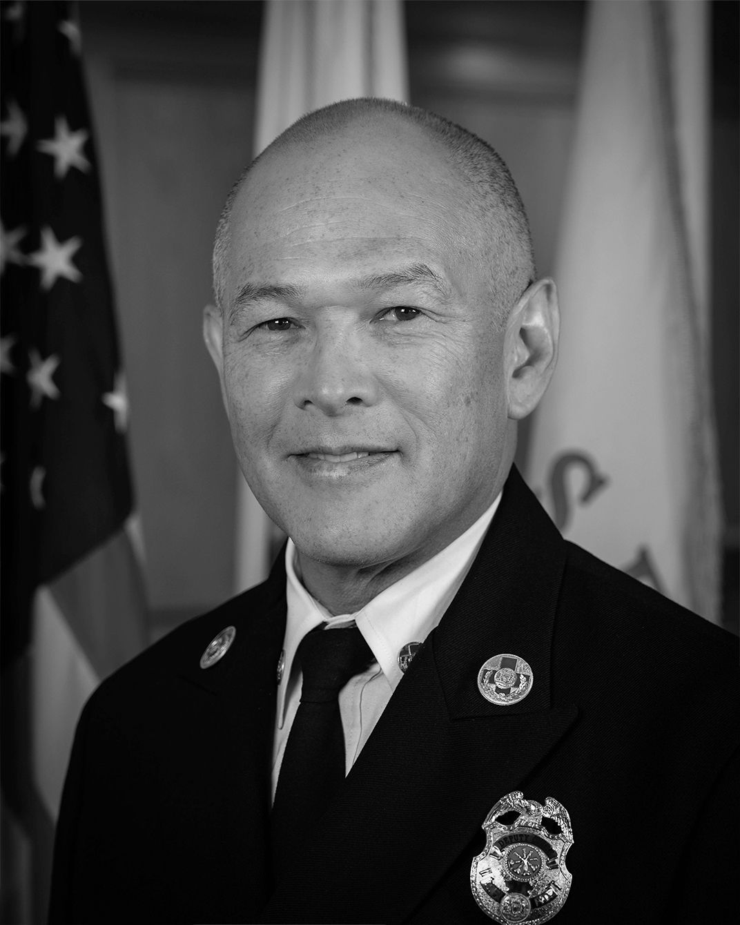 Formal portrait of a uniformed officer standing before U.S. and state flags, wearing a slight smile.