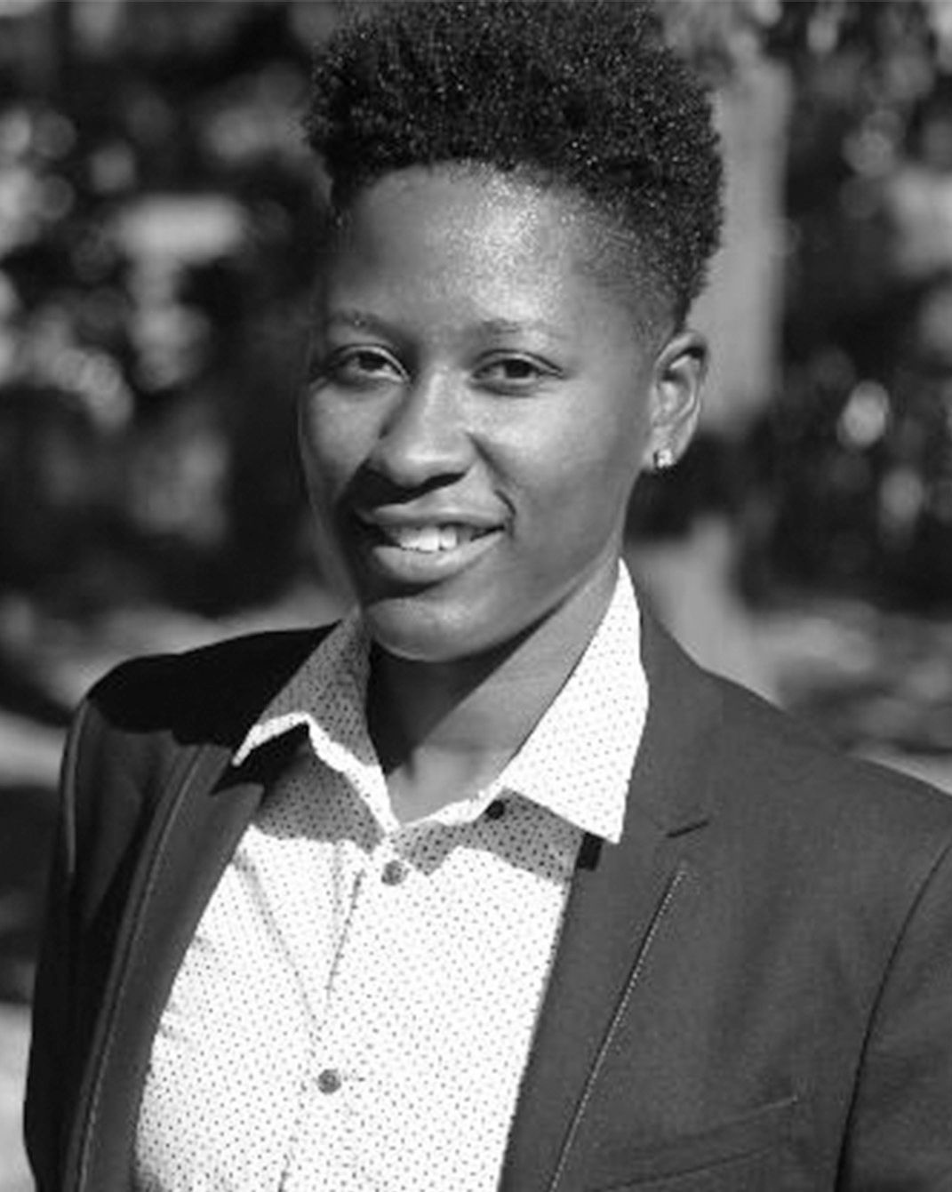 Smiling person in a blazer and patterned shirt, standing outdoors in black and white.