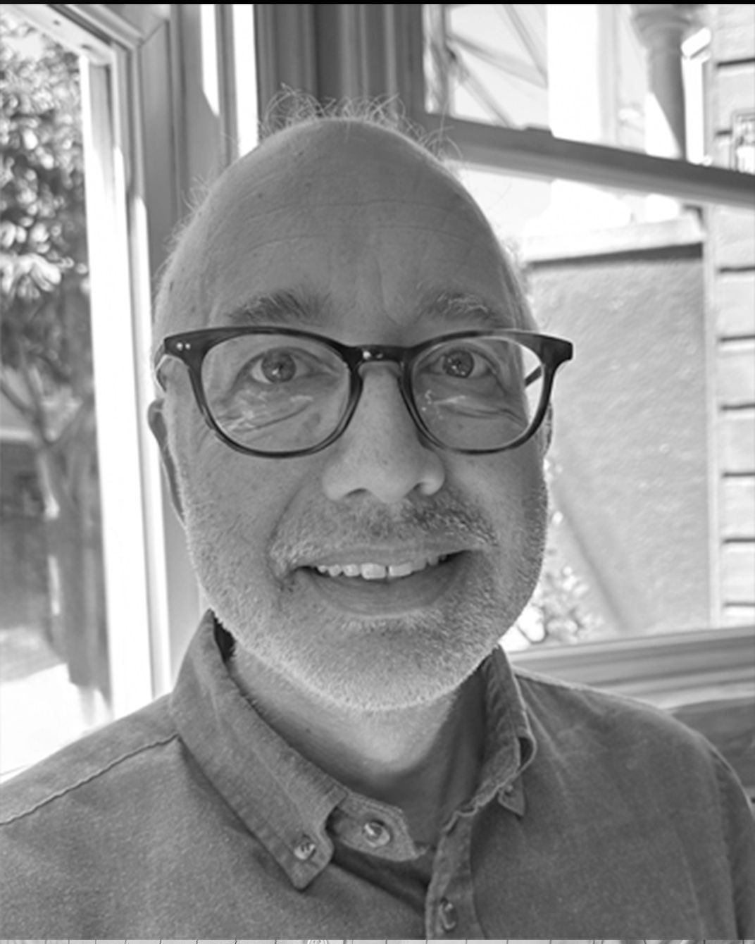 Black-and-white selfie of a smiling person wearing glasses near a window with curtains