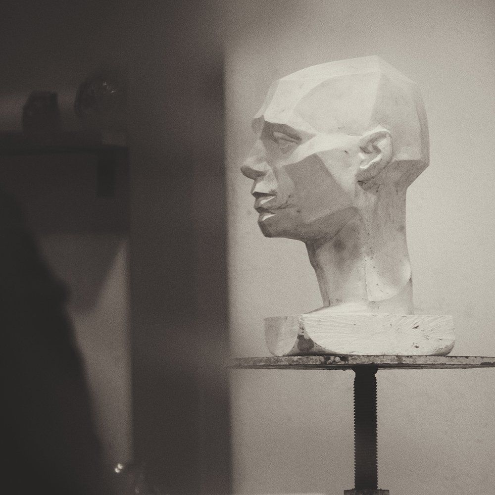 Monochrome bust sculpture in profile on a pedestal, lit against a plain indoor wall.