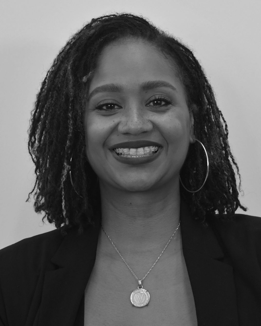 Smiling person with shoulder-length hair, hoop earrings, and a necklace, posing against a plain background.