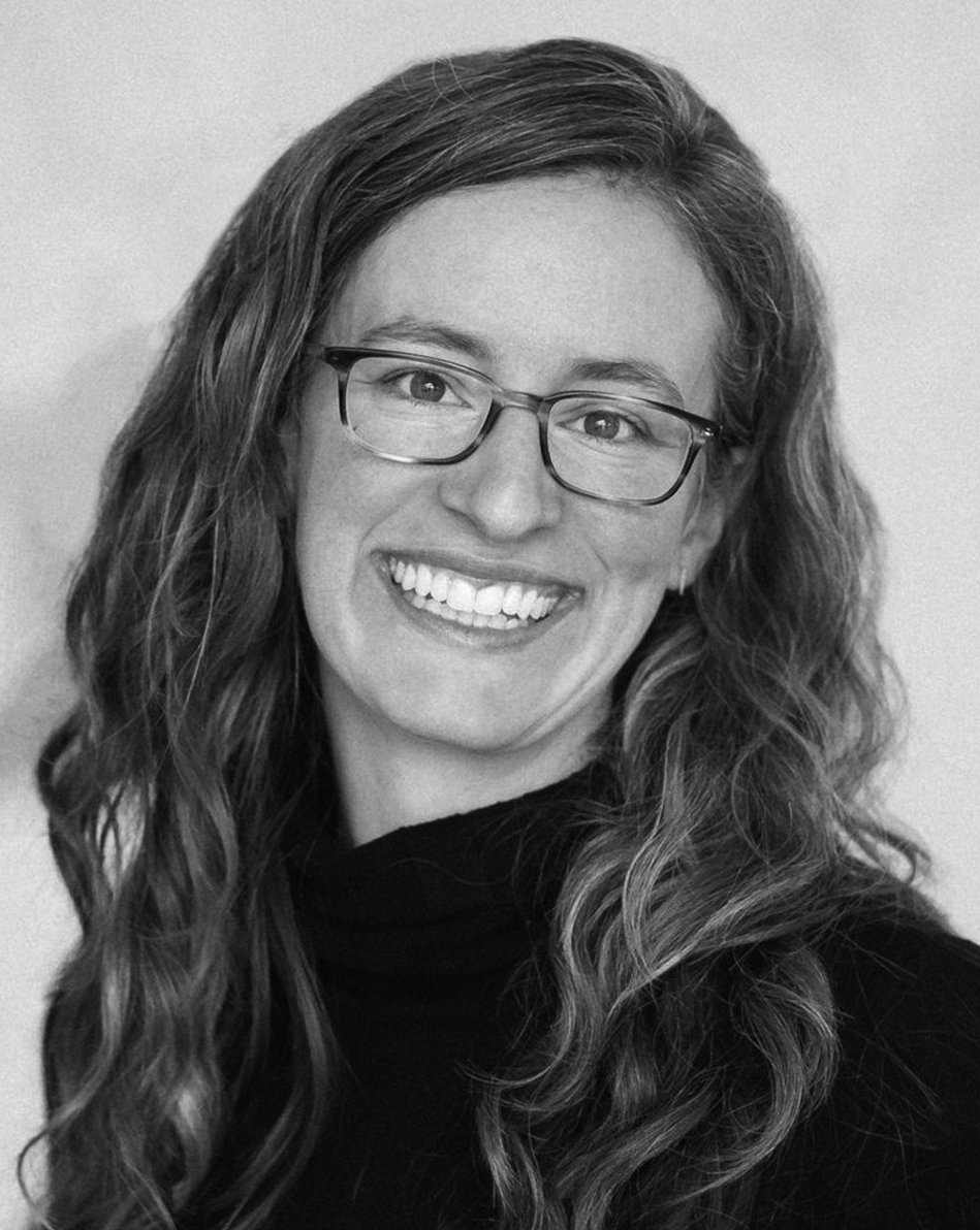 Smiling person with long wavy hair and glasses, wearing a dark top against a plain background