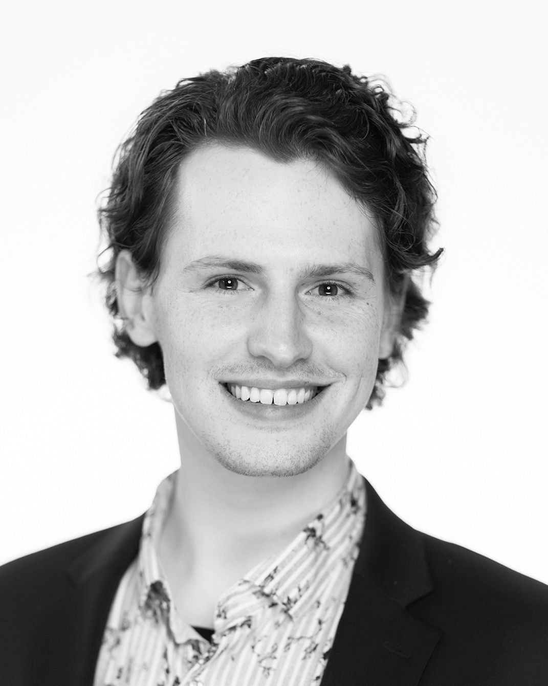 Smiling headshot of a person in a dark blazer and patterned shirt against a white background