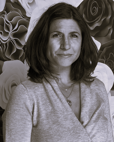 Portrait of a woman in a gray top standing before a floral mural, with a slight smile.