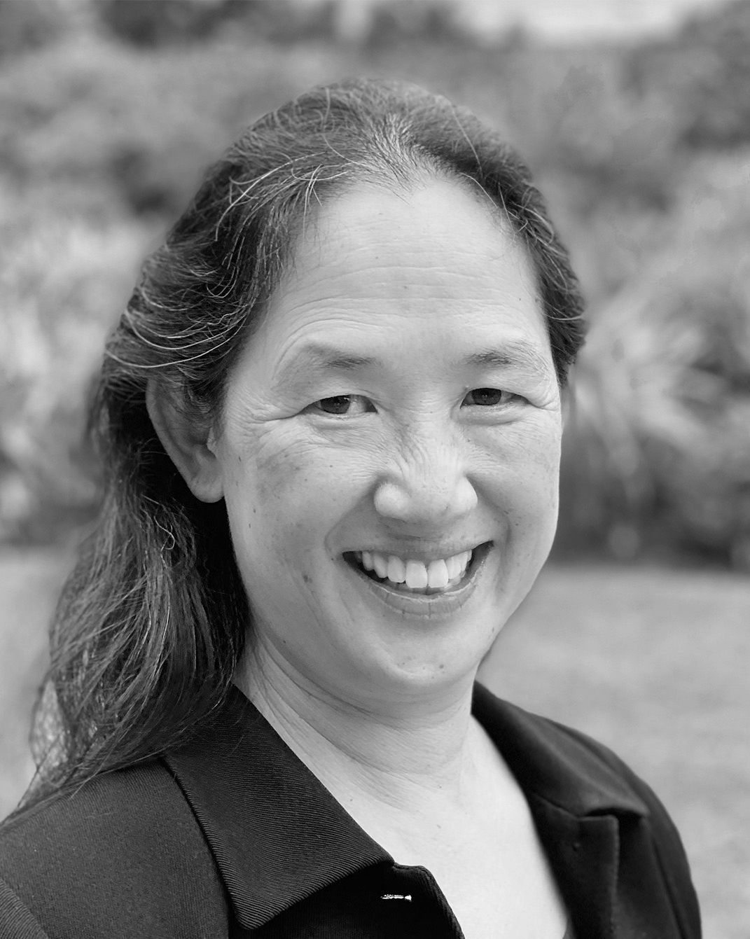 Smiling person with long hair in a black top, photographed in black and white outdoors.