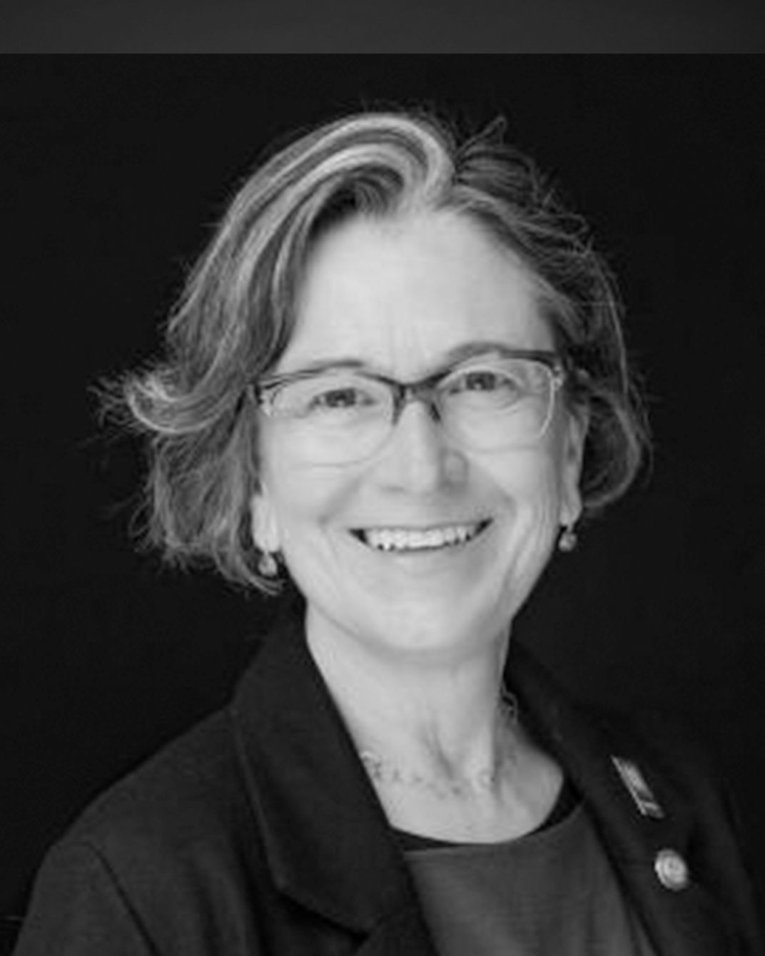 Smiling person in glasses and a dark blazer against a black background.