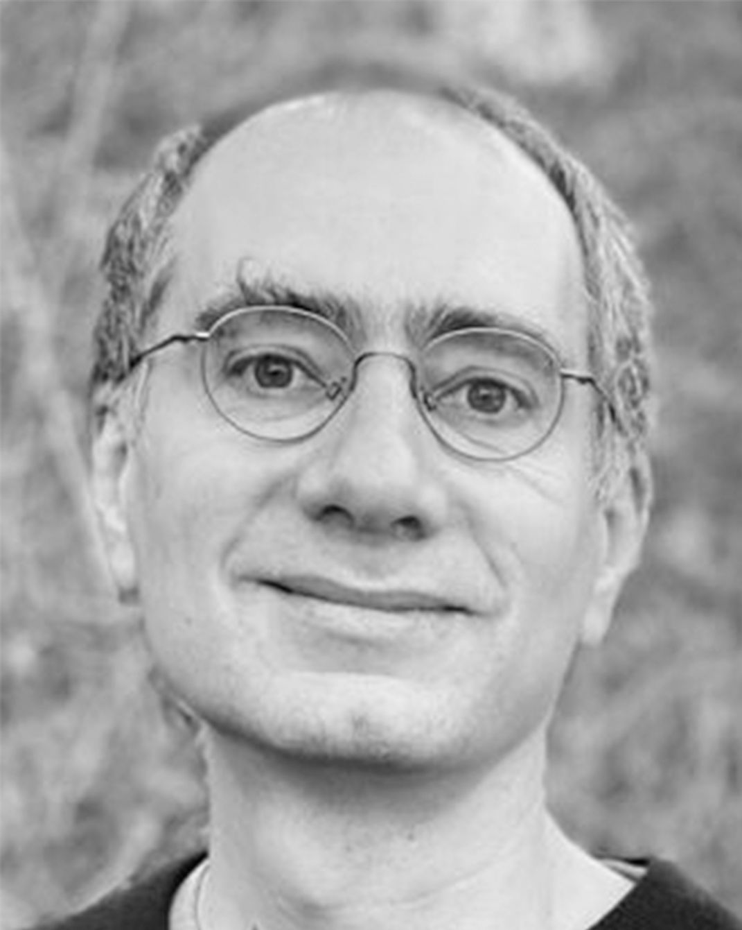 Black-and-white portrait of a smiling person wearing round glasses, outdoors with a blurred background