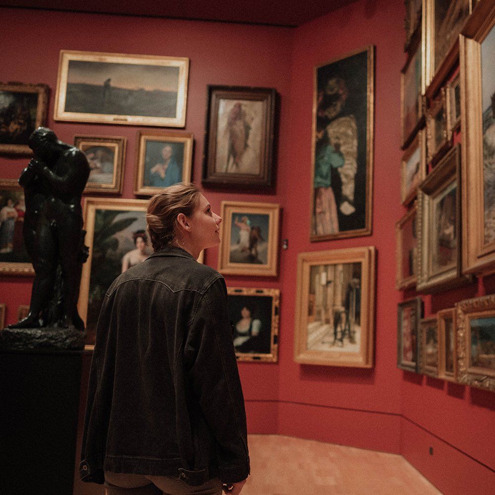 Person in a red-walled art gallery, viewing framed paintings and sculptures.