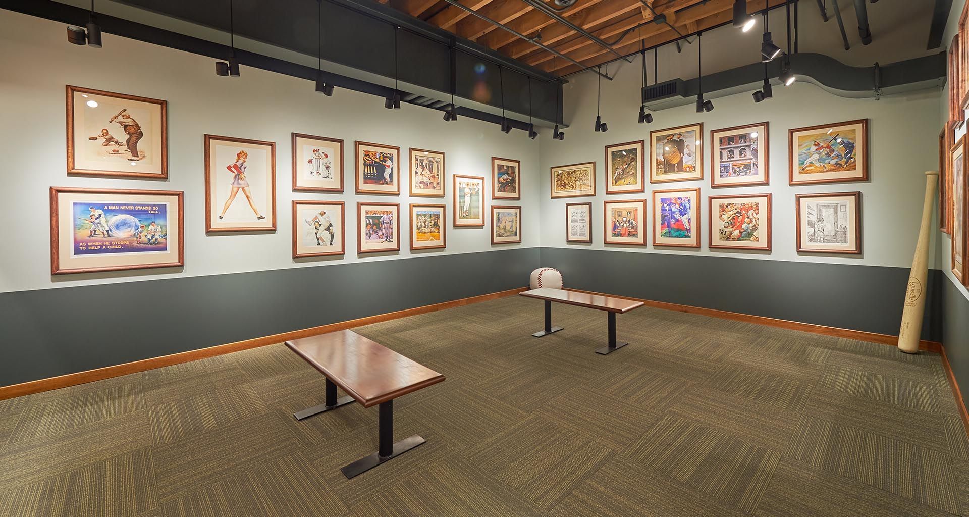 Art gallery room with framed artwork on gray walls, carpeted floor, and two benches under warm lighting