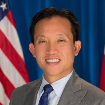 Portrait of a man in a gray suit and blue tie standing before U.S. and red flags