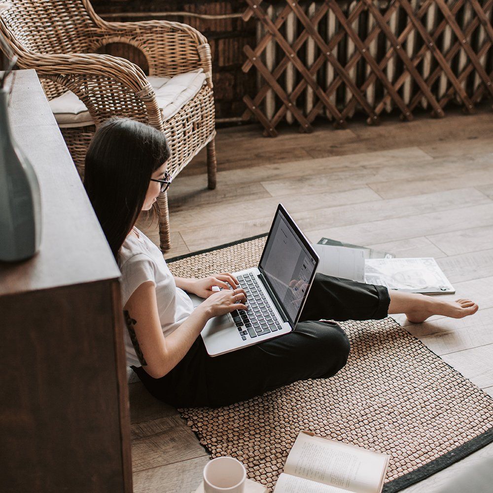 Person sitting on a rug using a laptop in a cozy room with wicker chairs and a coffee cup nearby