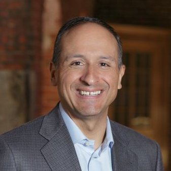 Smiling man in a gray blazer and light blue shirt against a warm indoor background