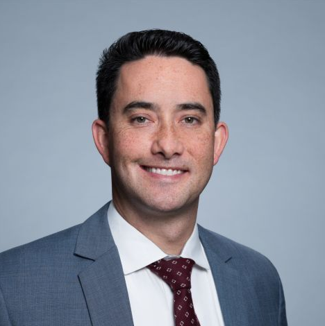 Professional headshot of a smiling man in a blue suit and maroon tie on a gray background