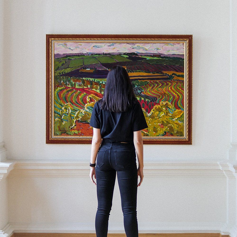 Woman stands facing a colorful landscape painting in a gallery.