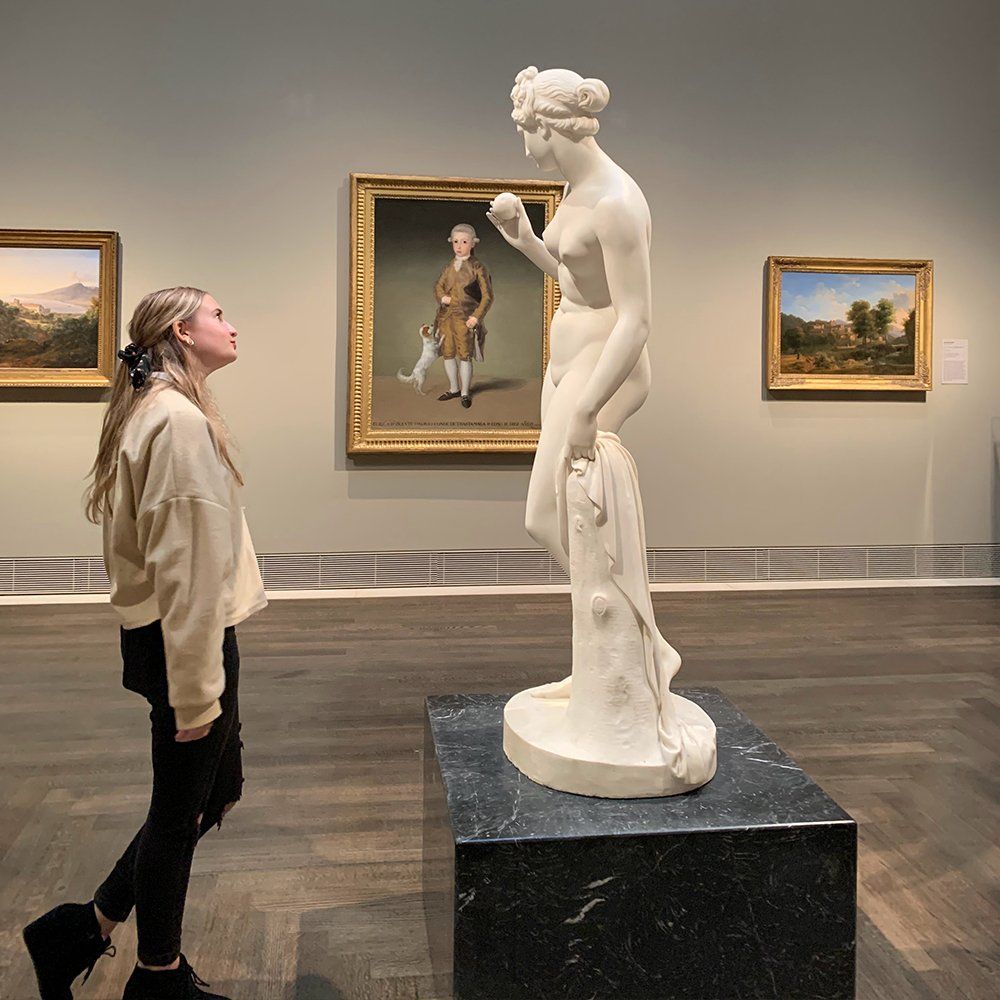 Girl looks at a white statue in an art gallery, with framed paintings on the walls.