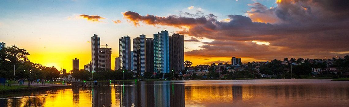 Vista da cidade ao pôr do sol, refletida em um lago tranquilo. Tons de laranja e dourado dominam o céu e a água.