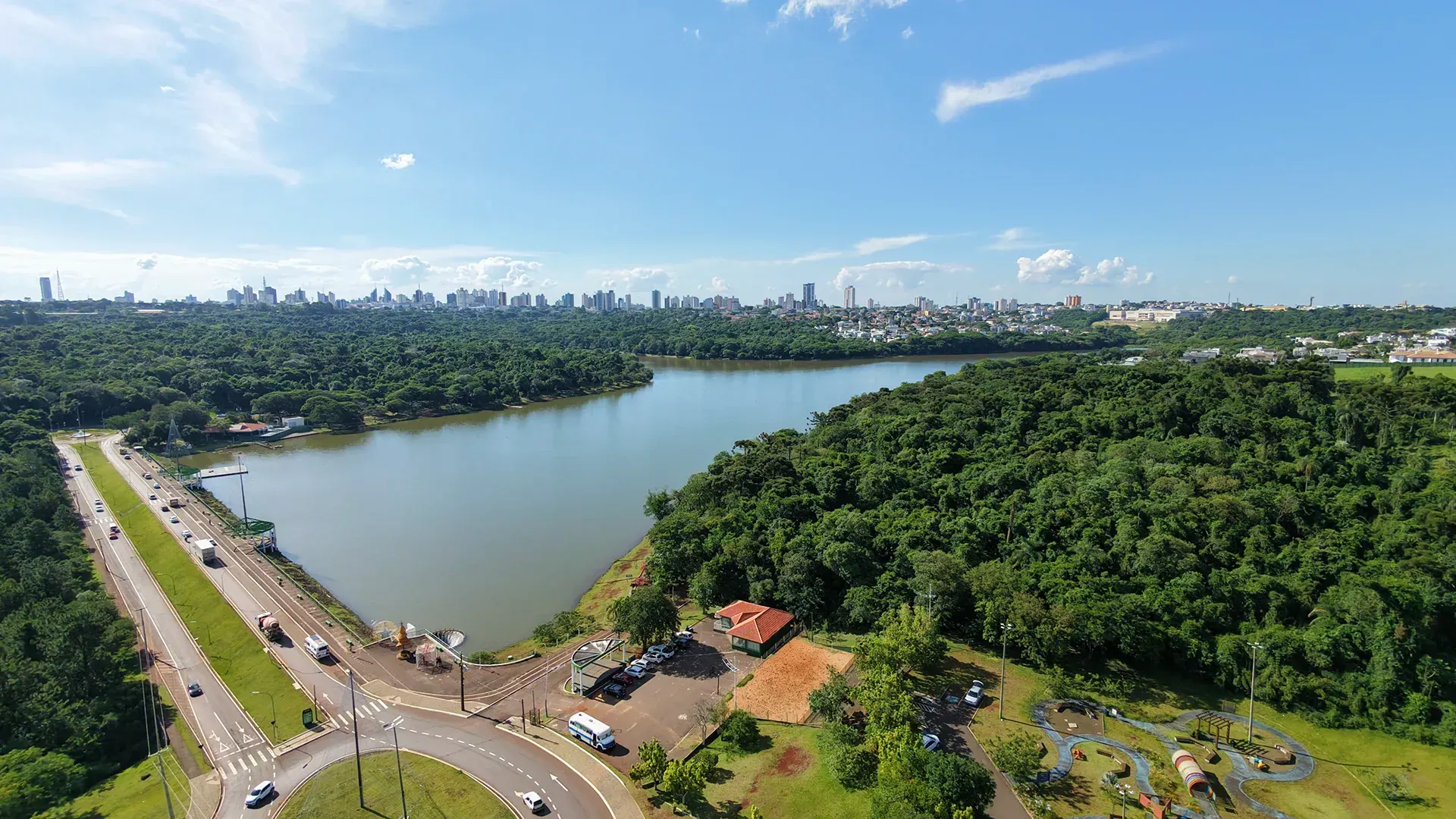 Ampla vista aérea de um lago e árvores verdes com o horizonte da cidade ao longe, sob um céu azul.