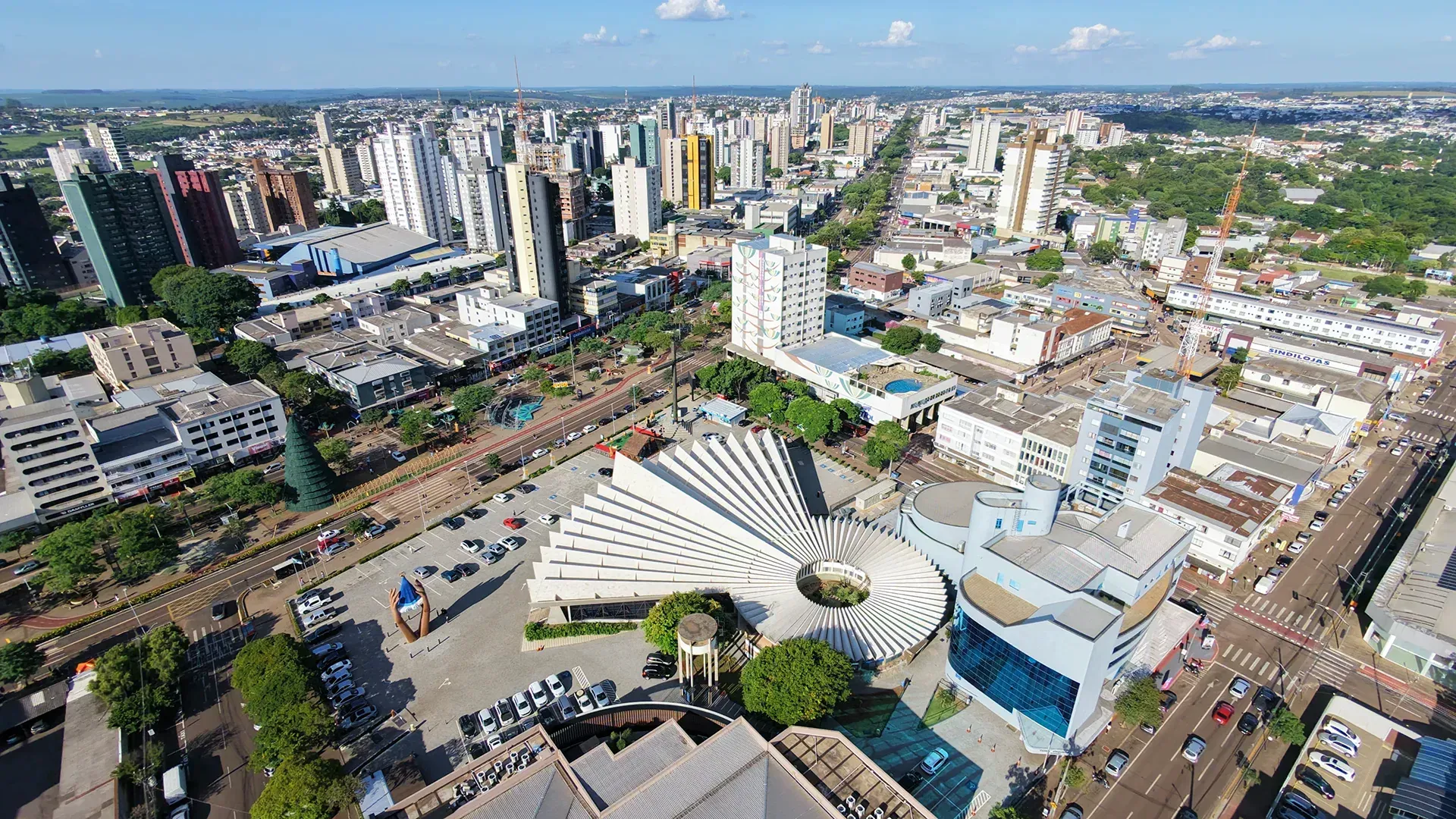 Vista aérea de uma cidade com prédios altos, um grande edifício moderno com telhado curvo e um amplo bulevar.