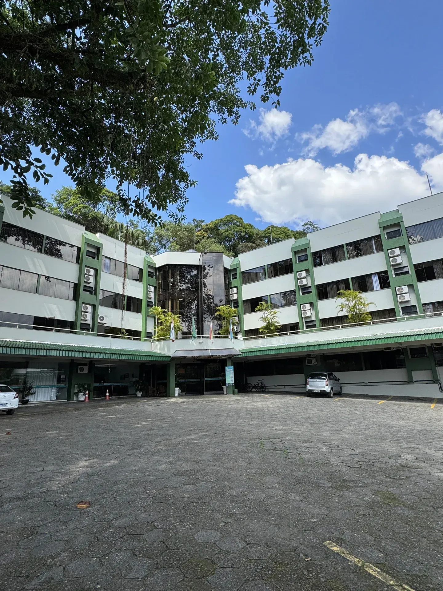 Prédio de vários andares, verde e branco, com janelas de vidro, situado em um estacionamento sob um céu azul com árvores.