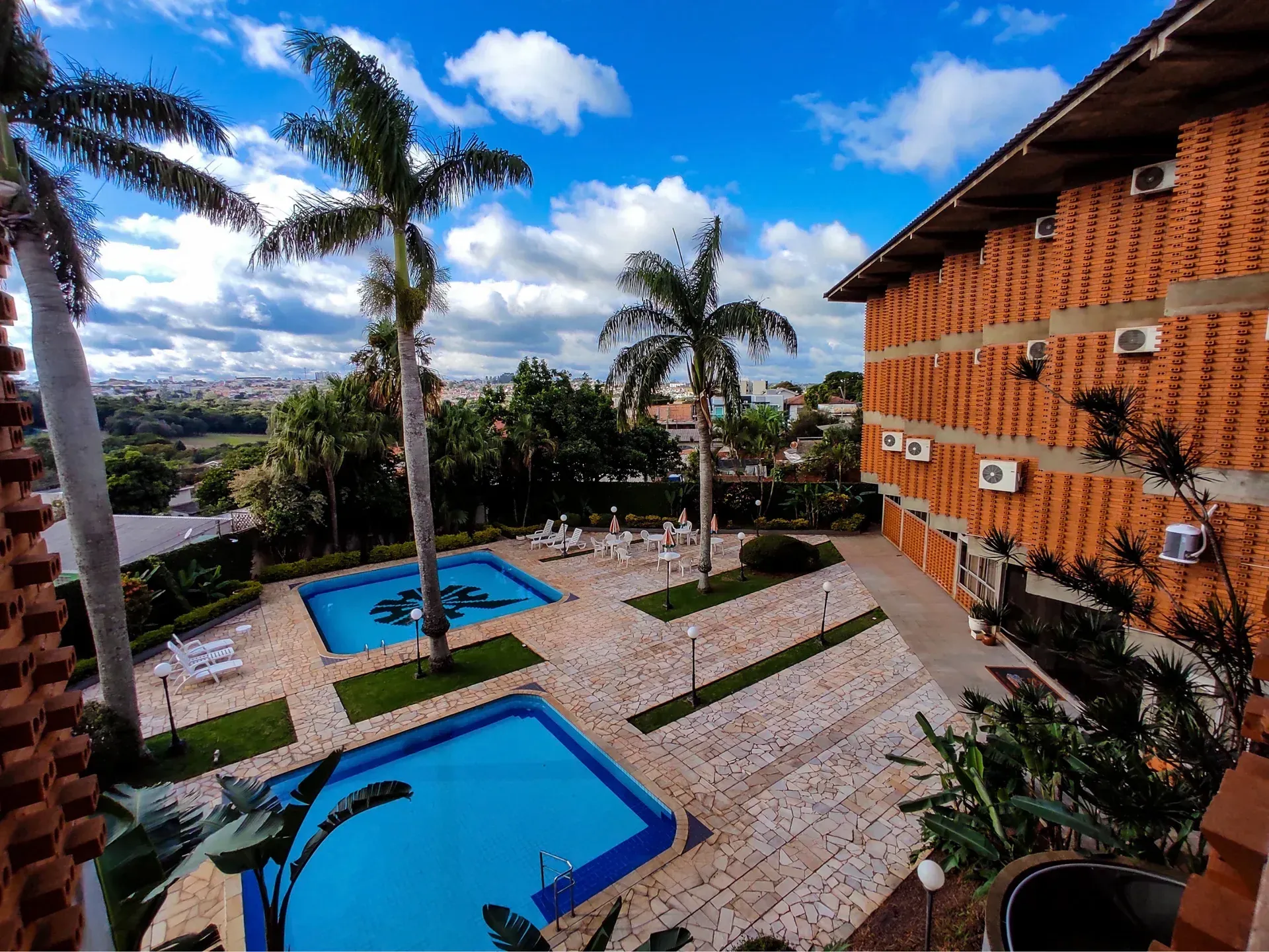 Vista exterior de um hotel com fachada de tijolos, duas piscinas, palmeiras e céu parcialmente nublado.