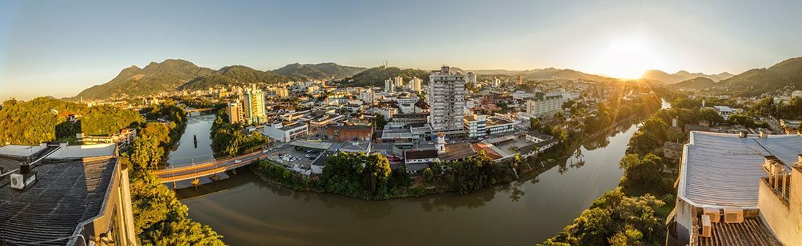 Vista aérea panorâmica de uma cidade às margens de um rio, com edifícios e montanhas visíveis ao pôr do sol.