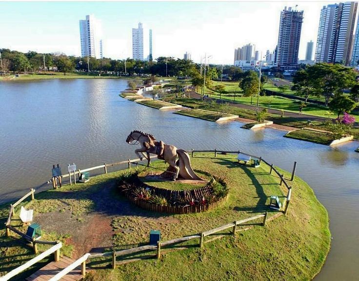 Uma estátua equestre de bronze em uma pequena ilha em um lago; horizonte da cidade ao fundo.