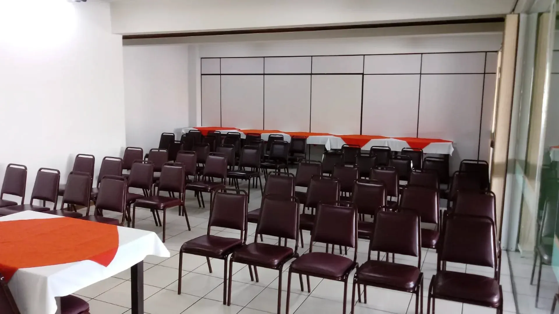 Sala de conferências vazia com fileiras de cadeiras, mesas e uma parede branca ao fundo.